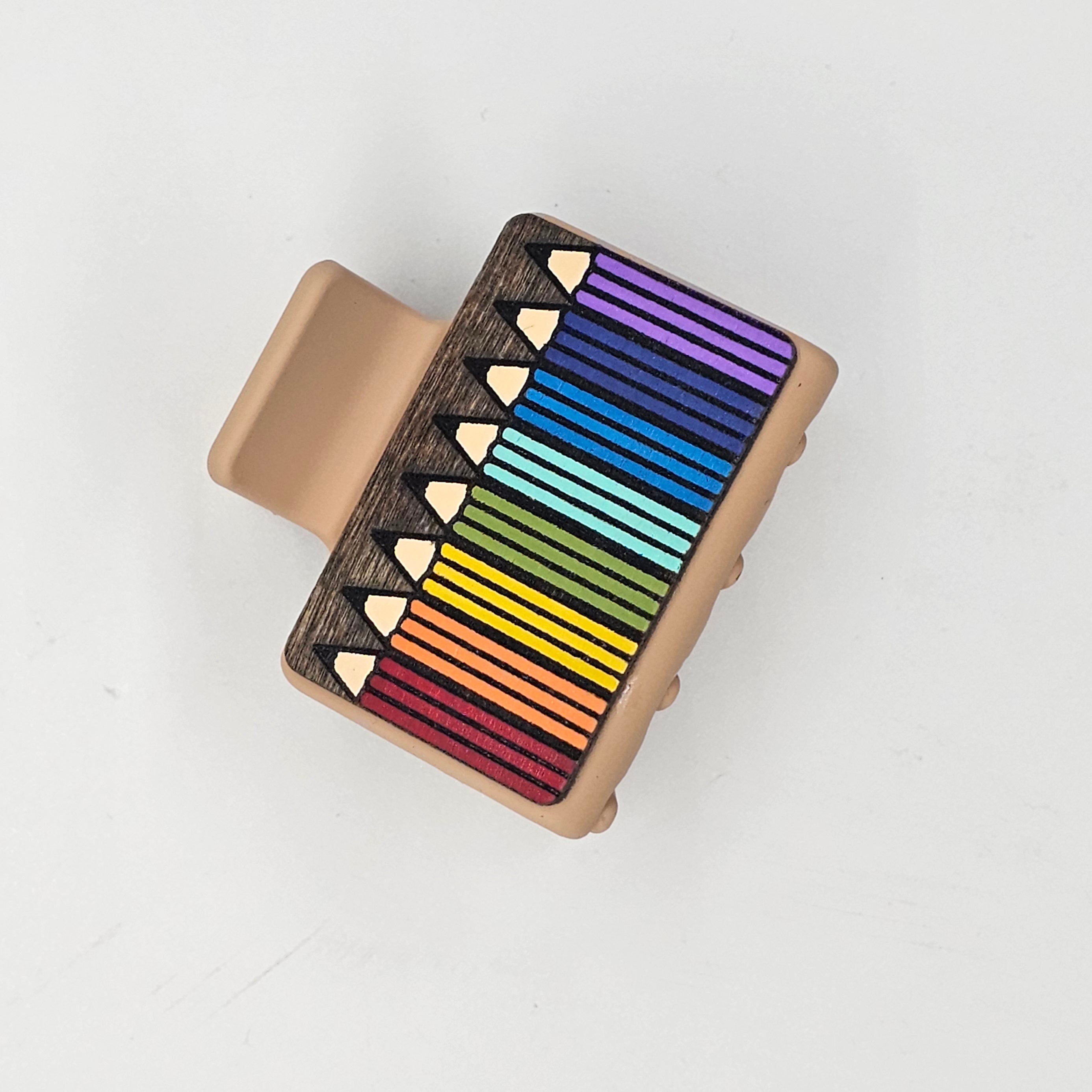 Hand-painted rainbow of colored pencils fanned diagonally with engraved natural wood tips on dark-stained wood, attached to a 2-inch matte tan claw clip