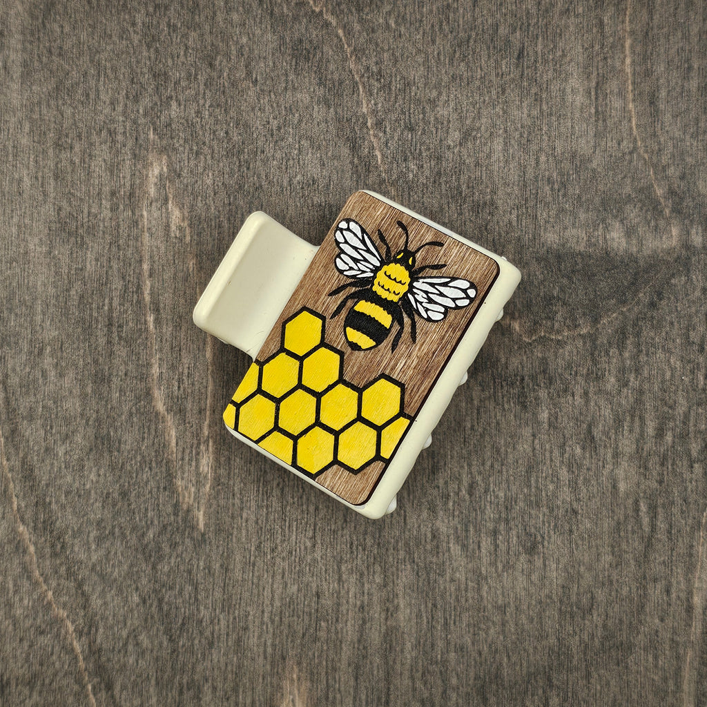 Hand-painted honeybee with engraved white wings and yellow-and-black striped body over bright yellow honeycomb grid on dark walnut wood grain, attached to a 2-inch matte cream claw clip