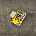 Hand-painted honeybee with engraved white wings and yellow-and-black striped body over bright yellow honeycomb grid on dark walnut wood grain, attached to a 2-inch matte cream claw clip