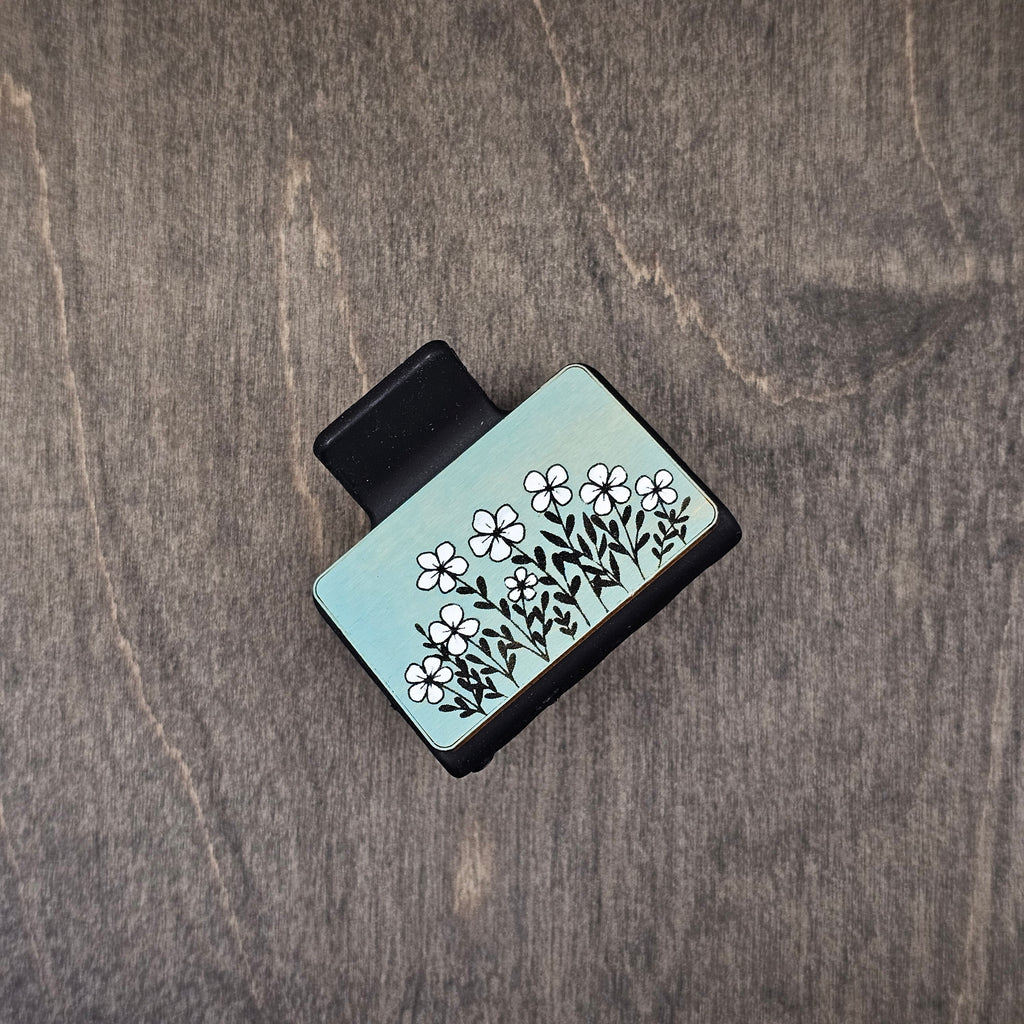 Hand-painted white wildflowers with engraved black stems and leaves on soft sage-mint blue wood tile, attached to a 2-inch matte black claw clip