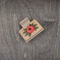 Hand-painted red poppy with engraved dark center and detailed green foliage on natural walnut wood grain, attached to a 2-inch matte taupe claw clip