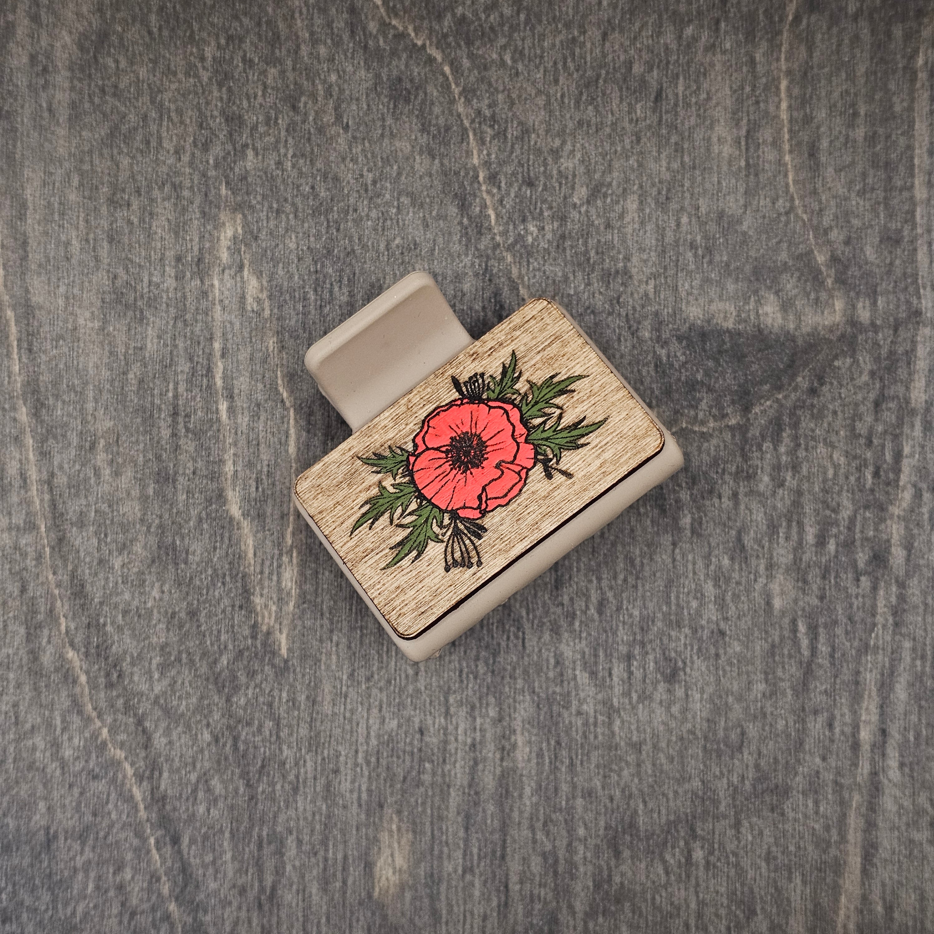 Hand-painted red poppy with engraved dark center and detailed green foliage on natural walnut wood grain, attached to a 2-inch matte taupe claw clip