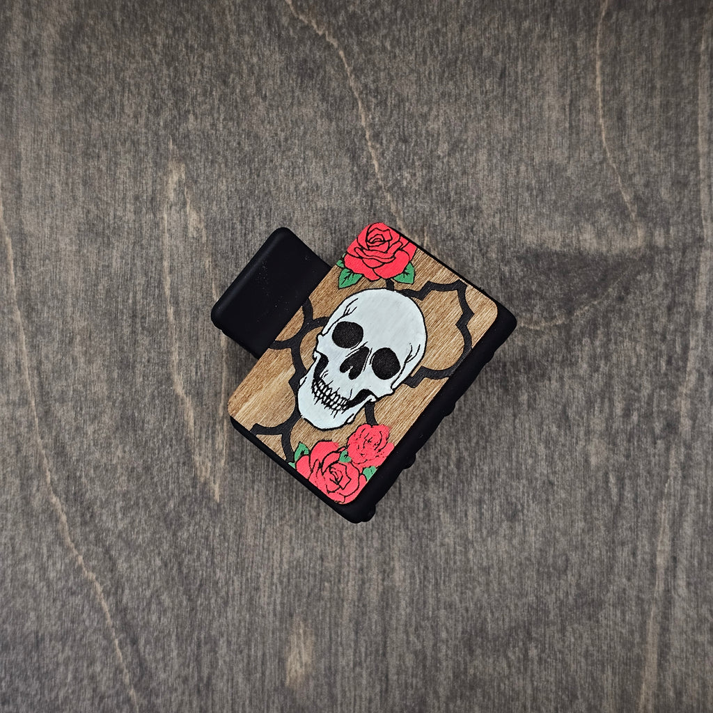 Hand-painted skull in white with black quatrefoil frame and vivid red roses with green leaves, laser engraved on natural walnut wood grain, attached to a 2-inch matte black claw clip