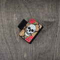 Hand-painted skull in white with black quatrefoil frame and vivid red roses with green leaves, laser engraved on natural walnut wood grain, attached to a 2-inch matte black claw clip