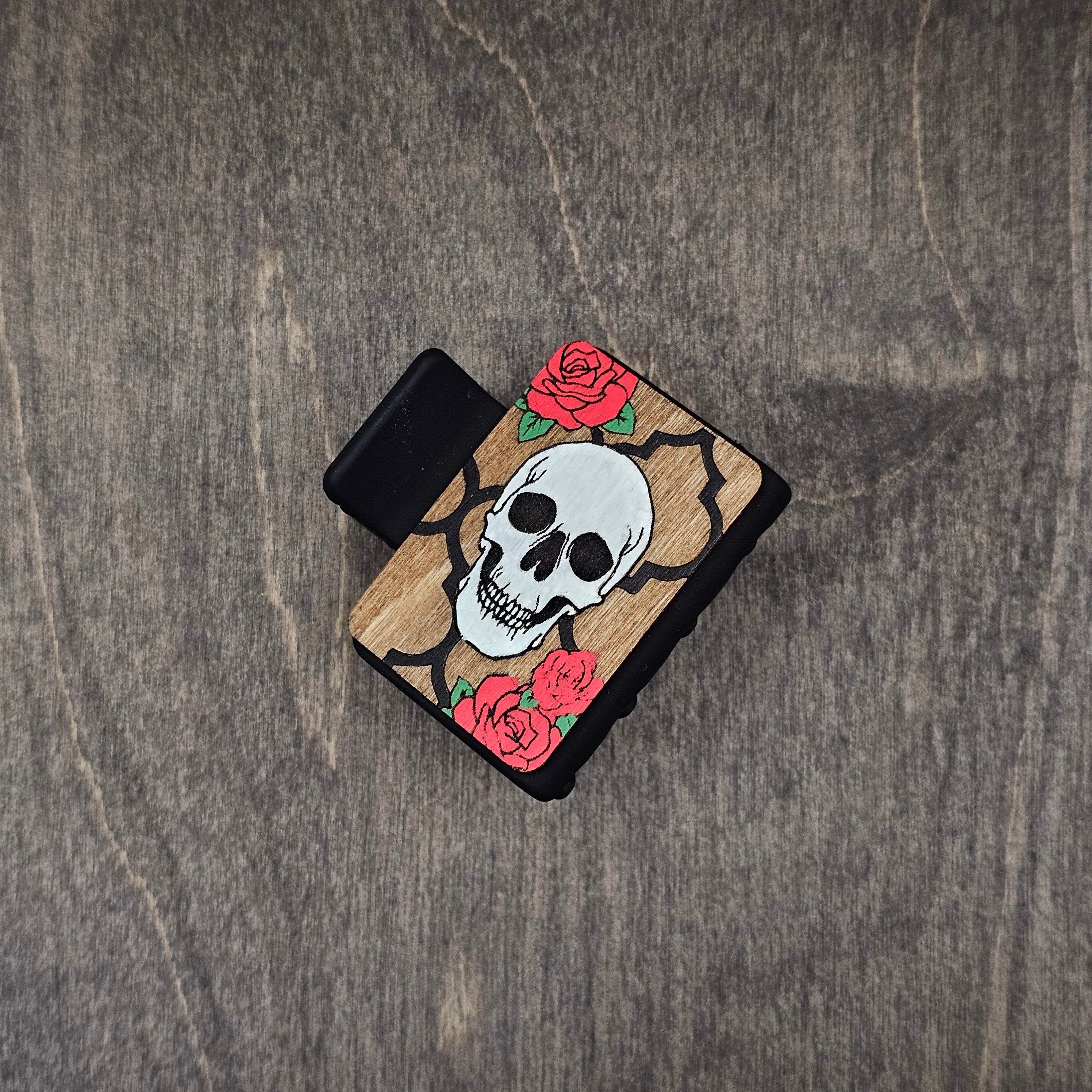 Hand-painted skull in white with black quatrefoil frame and vivid red roses with green leaves, laser engraved on natural walnut wood grain, attached to a 2-inch matte black claw clip