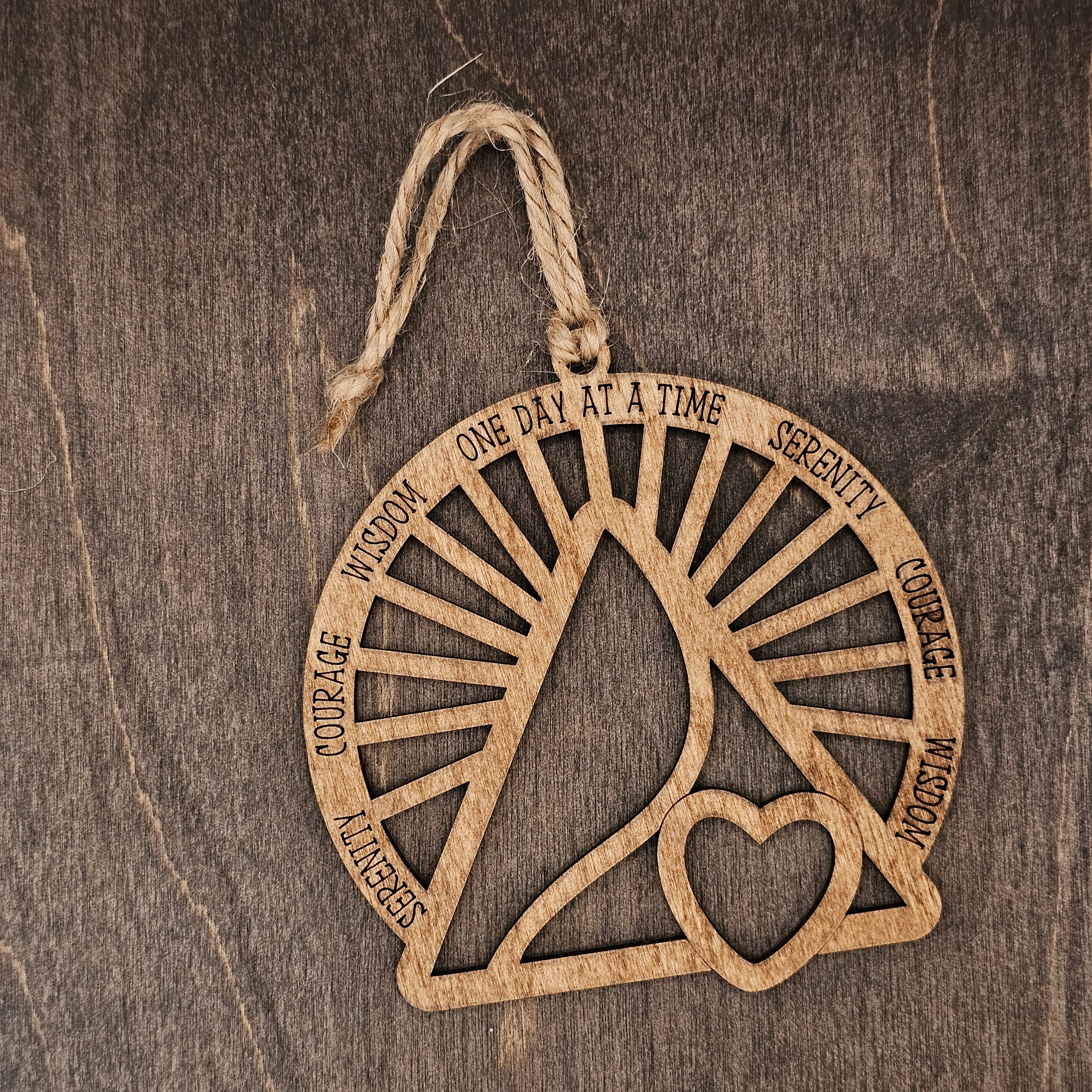 Laser cut wood recovery ornament with open sunburst design engraved with Serenity, Courage, Wisdom, and One Day at a Time, with a heart cutout at the center, hung with natural jute cord
