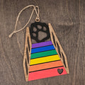 Laser cut Rainbow Bridge dog ornament with hand-painted rainbow colored steps, black dog paw print silhouette, and heart cutout at the base, hung with natural jute cord