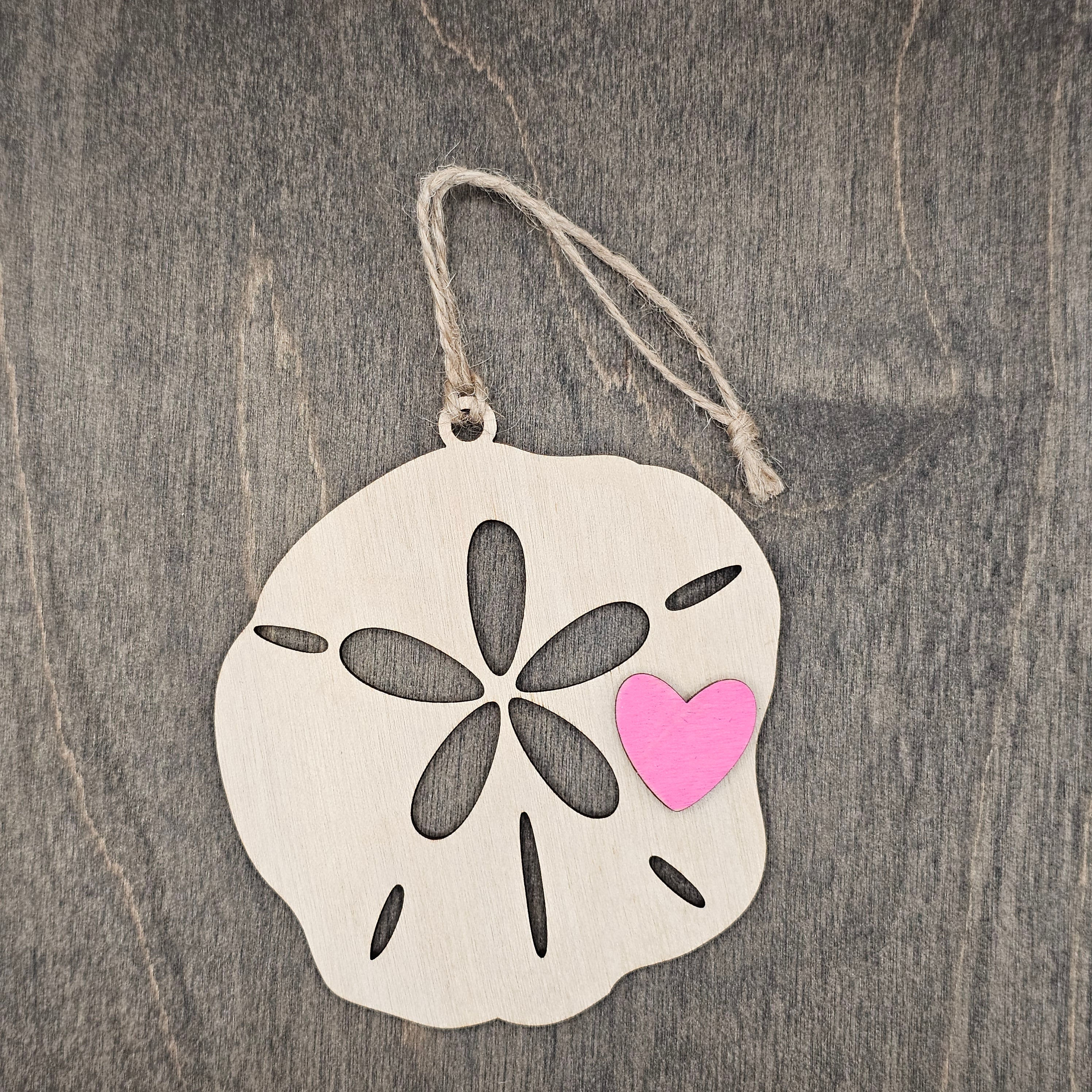 Laser cut wood sand dollar ornament with five-petal cutout and hand-painted pink heart accent, hung with natural jute cord