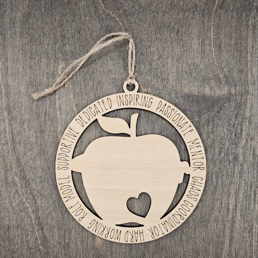 Laser cut wood apple ornament with heart cutout inside a circular frame engraved with teacher descriptors including Dedicated, Inspiring, Passionate, Mentor, Chaos Coordinator, Hard Working, Role Model, and Supportive, hung with natural jute cord
