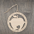 Laser cut wood apple ornament with heart cutout inside a circular frame engraved with teacher descriptors including Dedicated, Inspiring, Passionate, Mentor, Chaos Coordinator, Hard Working, Role Model, and Supportive, hung with natural jute cord