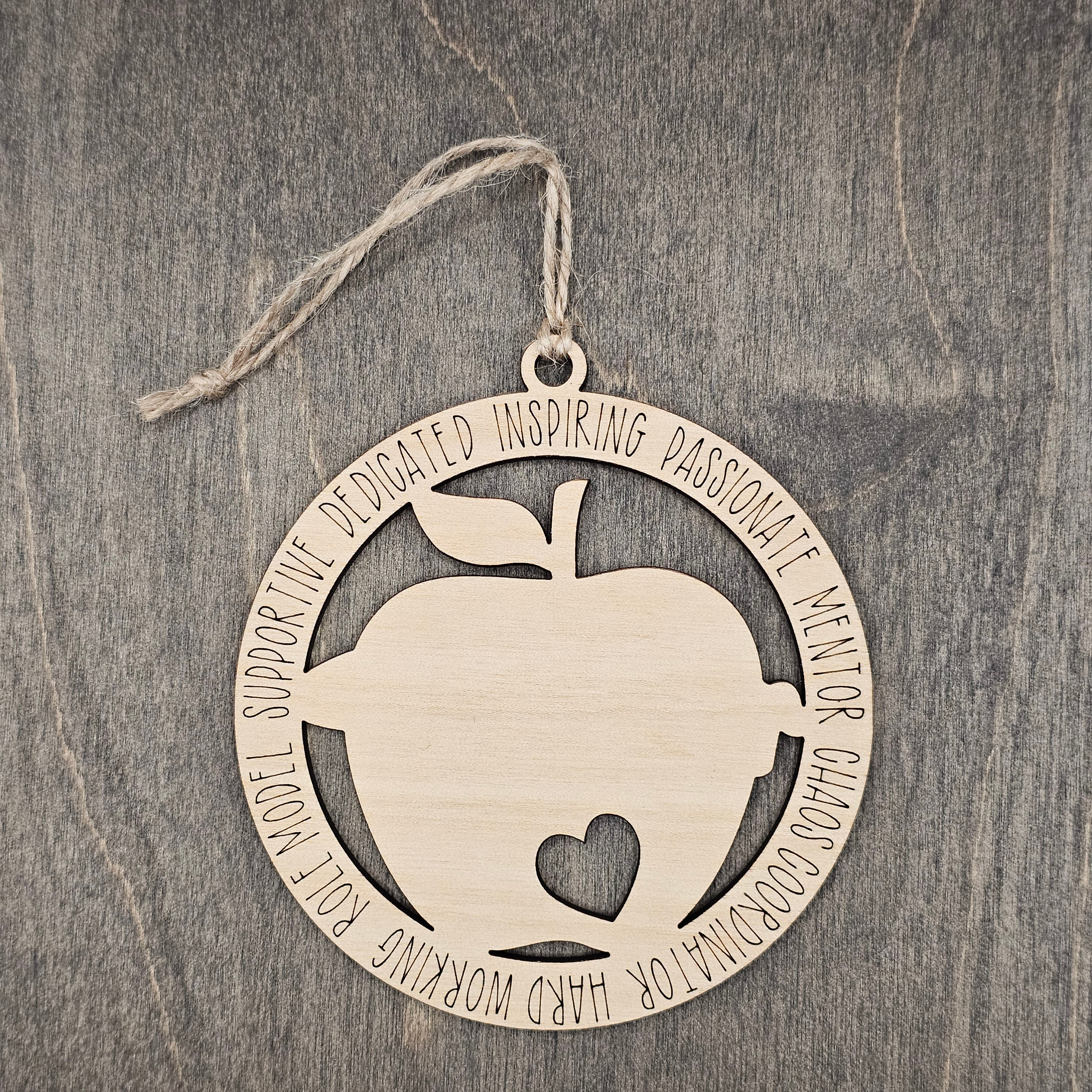 Laser cut wood apple ornament with heart cutout inside a circular frame engraved with teacher descriptors including Dedicated, Inspiring, Passionate, Mentor, Chaos Coordinator, Hard Working, Role Model, and Supportive, hung with natural jute cord