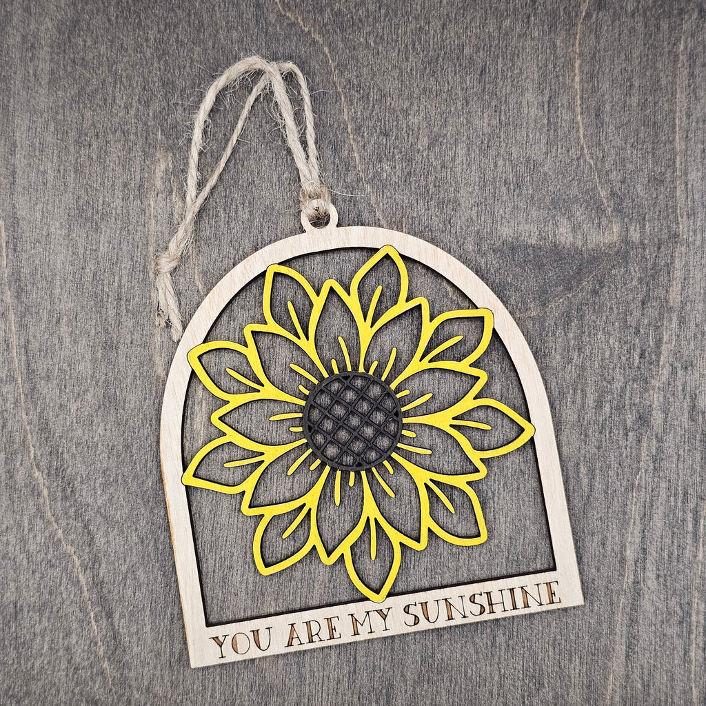 Hand-painted laser cut sunflower wood ornament in yellow with dark brown center inside an arched frame, "You Are My Sunshine" engraved at the base, hung with natural jute cord