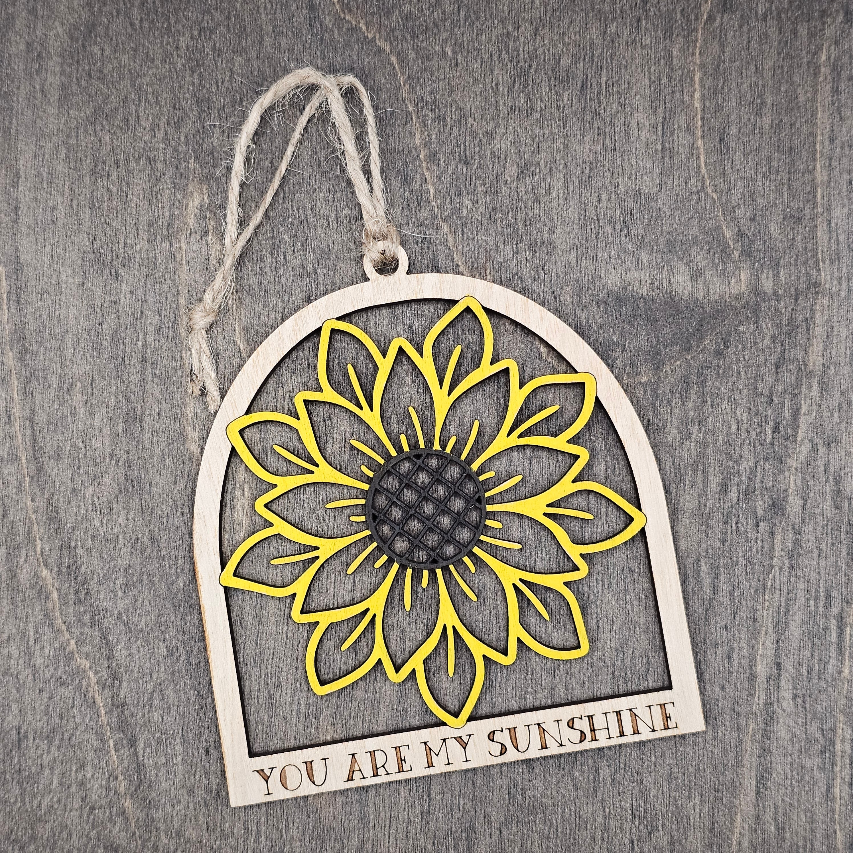 Hand-painted laser cut sunflower wood ornament in yellow with dark brown center inside an arched frame, "You Are My Sunshine" engraved at the base, hung with natural jute cord