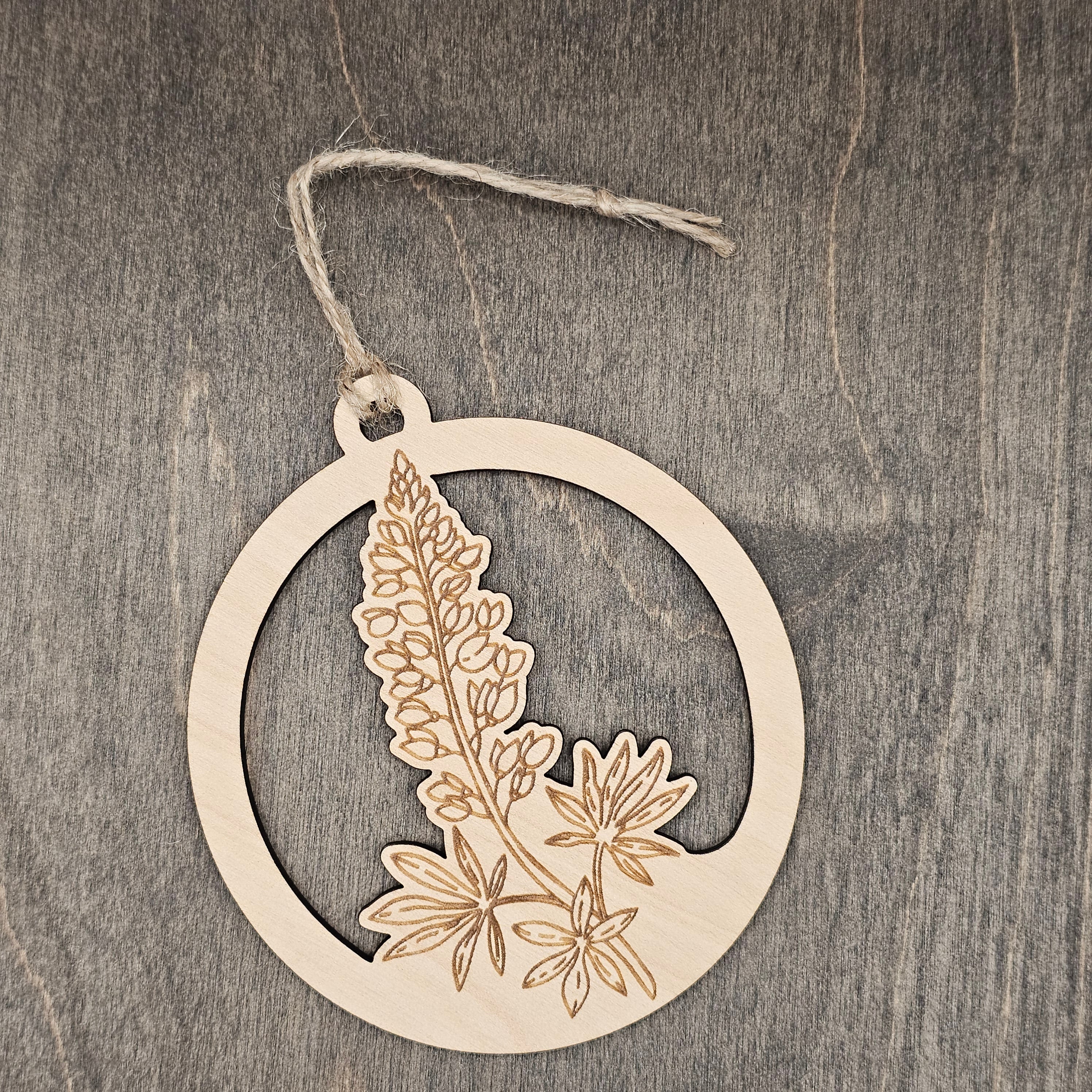 Laser cut and engraved Texas Bluebonnet wood ornament, featuring a delicate open silhouette floral design inside a circular wood frame, hung with natural jute cord