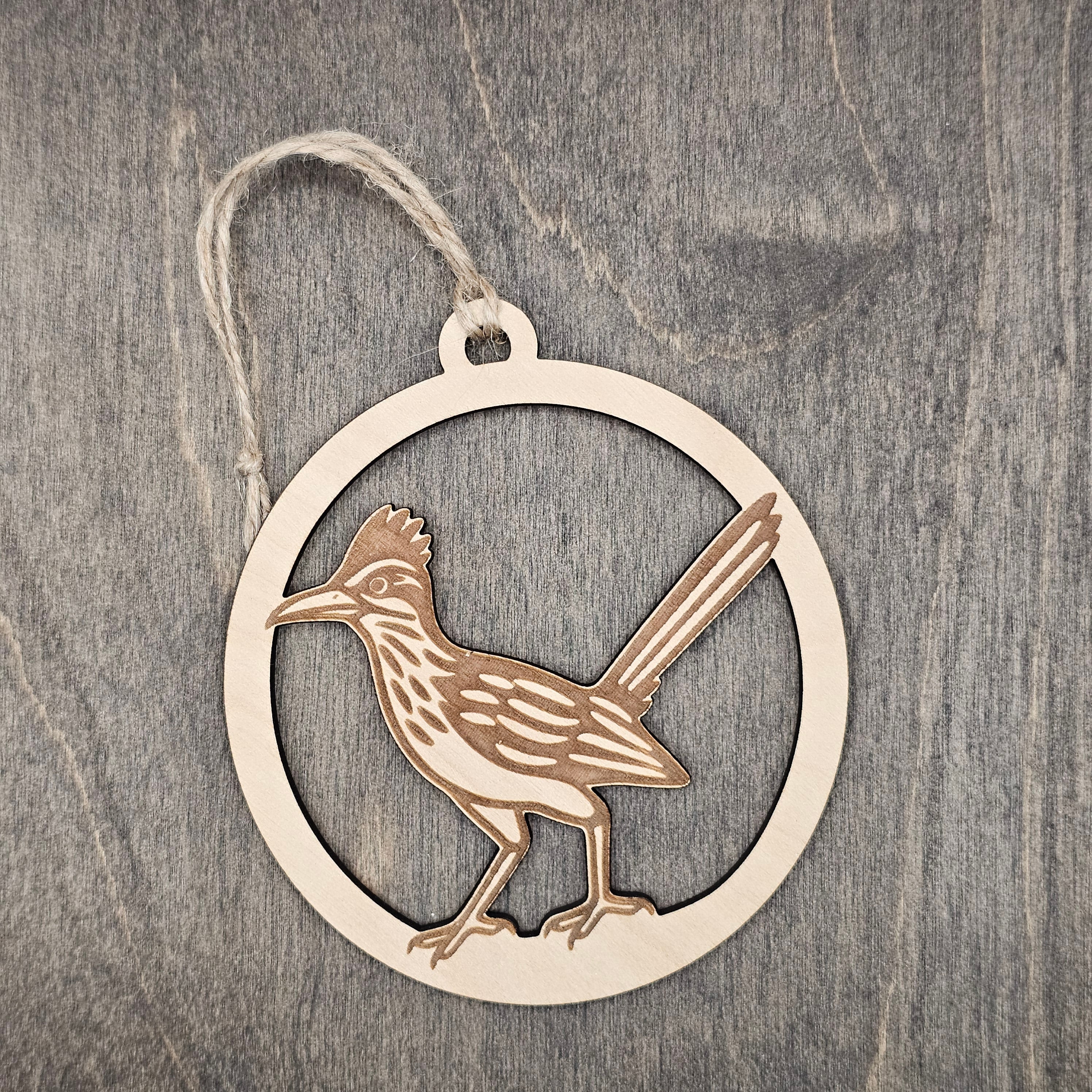 Laser cut and engraved Greater Roadrunner wood ornament, featuring an open silhouette design with detailed feather engraving inside a circular wood frame, hung with natural jute cord
