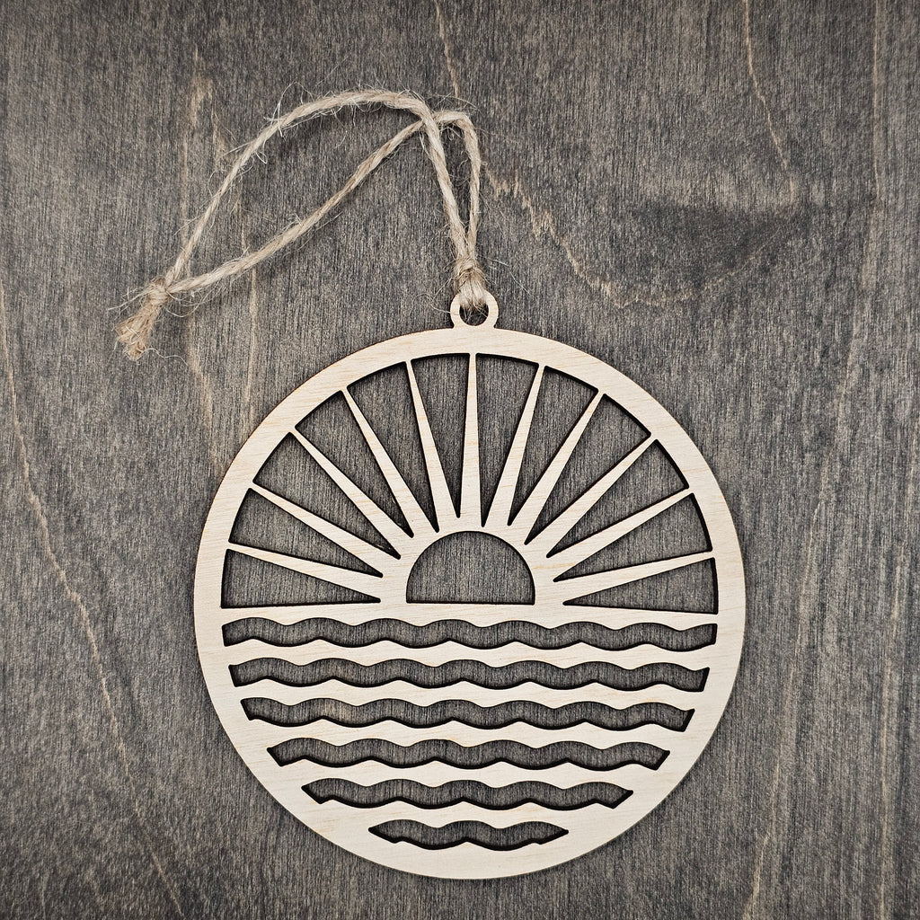 Laser cut wood sunrise ornament with radiating sun rays and rolling waves inside a circular frame, hung with natural jute cord