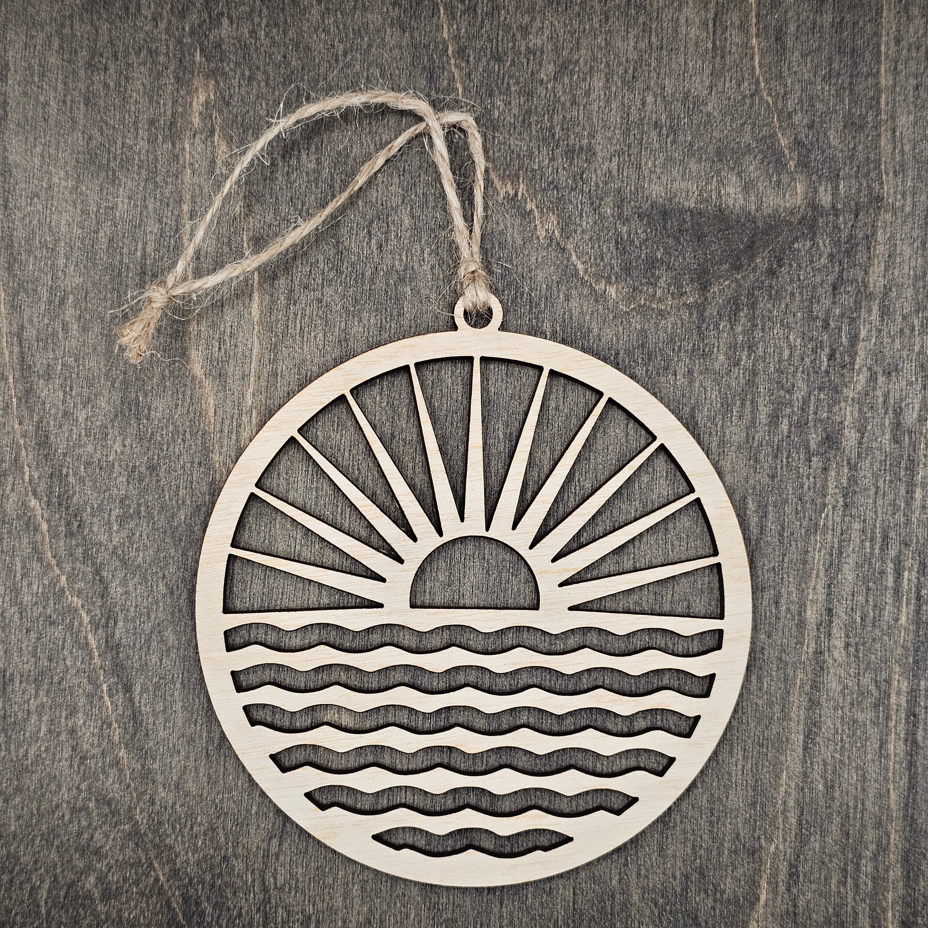 Laser cut wood sunrise ornament with radiating sun rays and rolling waves inside a circular frame, hung with natural jute cord