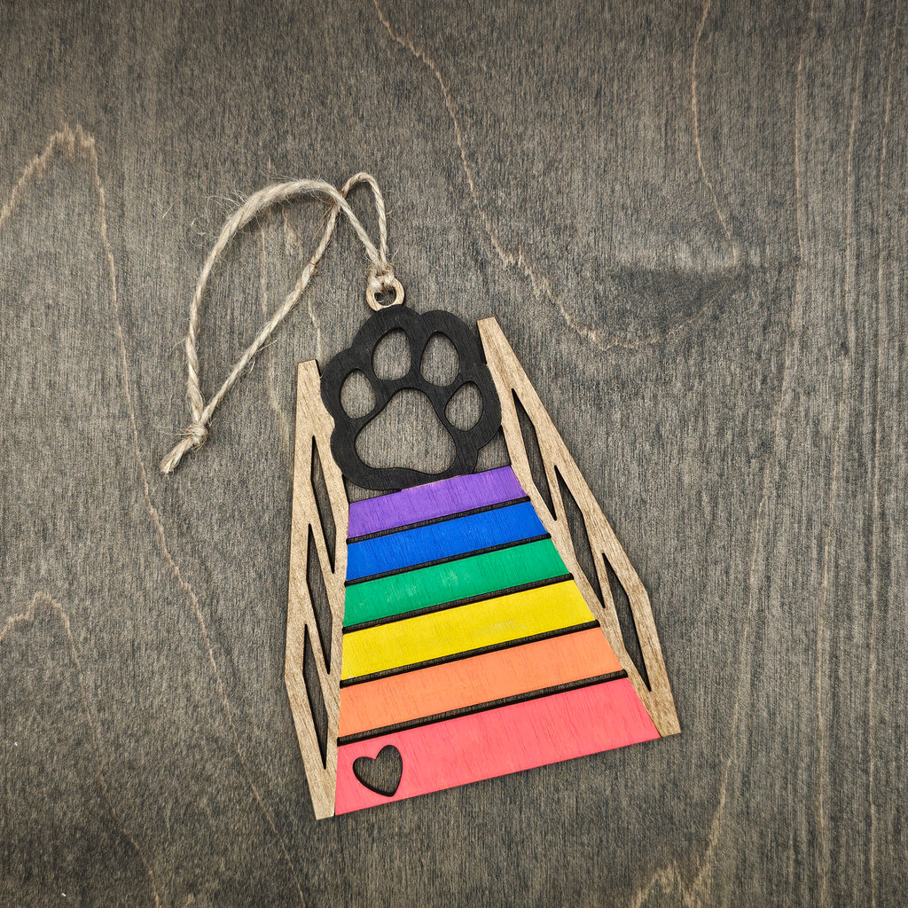 Laser cut Rainbow Bridge cat ornament with hand-painted rainbow colored steps, black cat paw print silhouette, and heart cutout at the base, hung with natural jute cord
