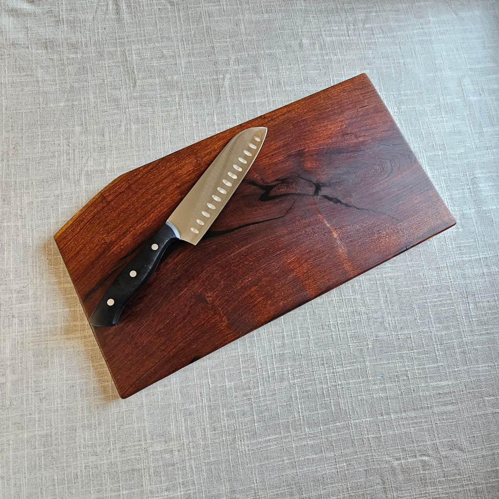 A rectangular dark brown wooden cutting board with a black handle and a Santoku knife, placed diagonally on a textured light gray linen tablecloth.
