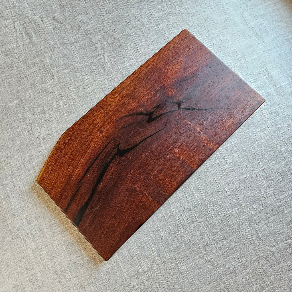 A rectangular wooden serving board with a natural live edge and prominent dark wood grain, displayed on a textured beige linen surface.