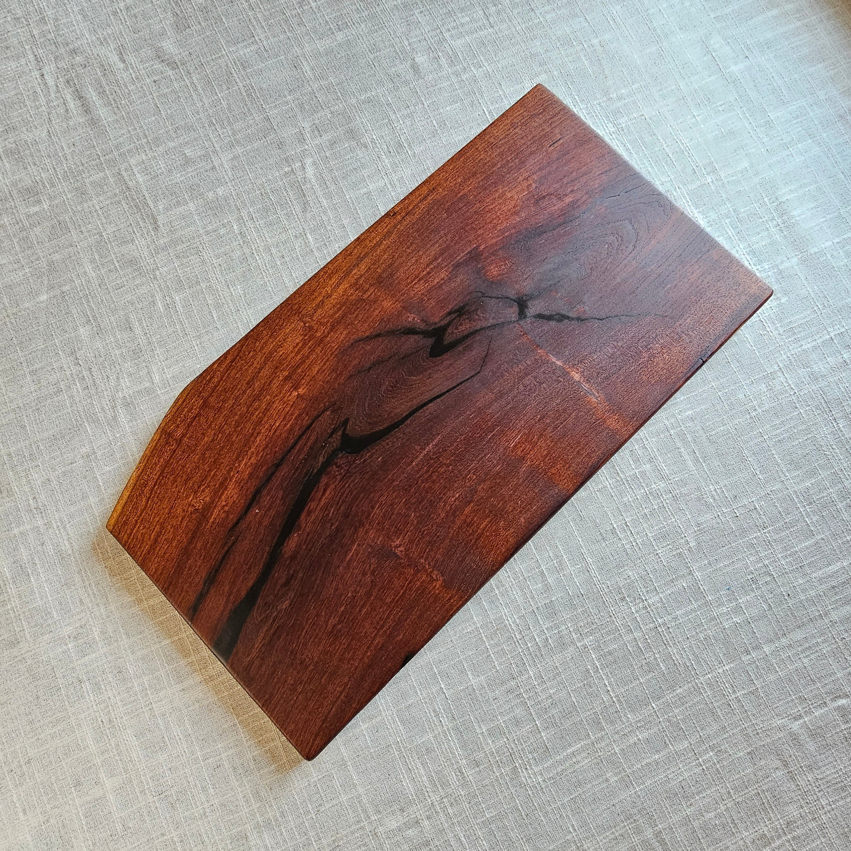 A rectangular wooden serving board with a natural live edge and prominent dark wood grain, displayed on a textured beige linen surface.