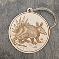 Laser engraved Armadillo wood ornament, featuring a detailed illustration of this Texas icon with its banded shell, walking through native grassland with yucca in the background, hung with natural jute cord
