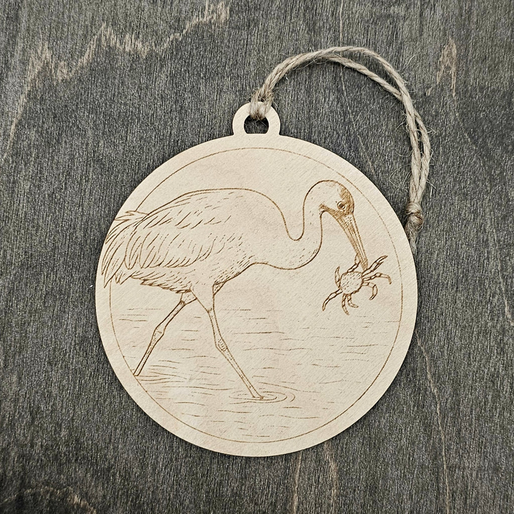 Laser engraved Whooping Crane wood ornament, featuring a detailed illustration of this endangered bird wading in coastal marsh waters with a blue crab, hung with natural jute cord