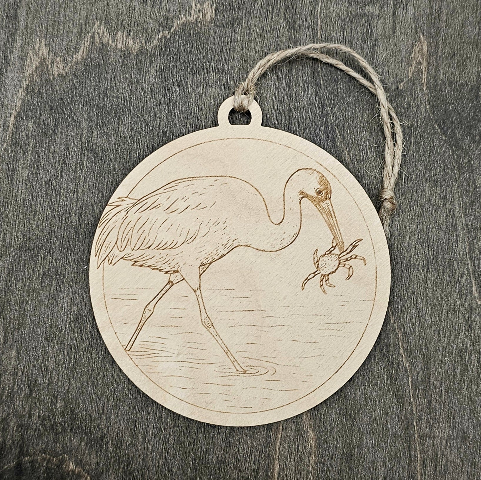 Laser engraved Whooping Crane wood ornament, featuring a detailed illustration of this endangered bird wading in coastal marsh waters with a blue crab, hung with natural jute cord