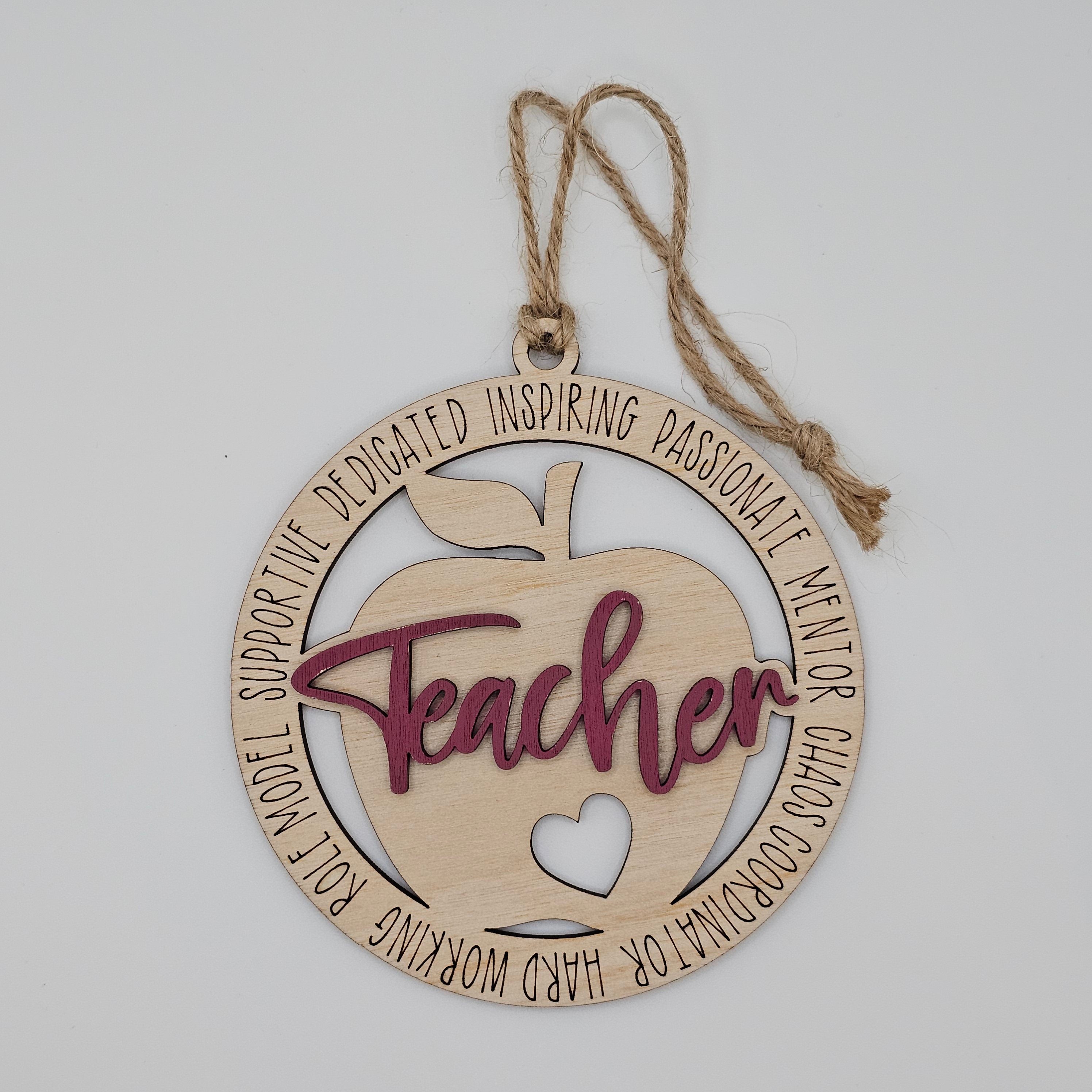 Layered laser cut Teacher apple wood ornament with maroon painted script overlay, circular frame engraved with teacher descriptors, heart cutout at center, hung with natural jute cord