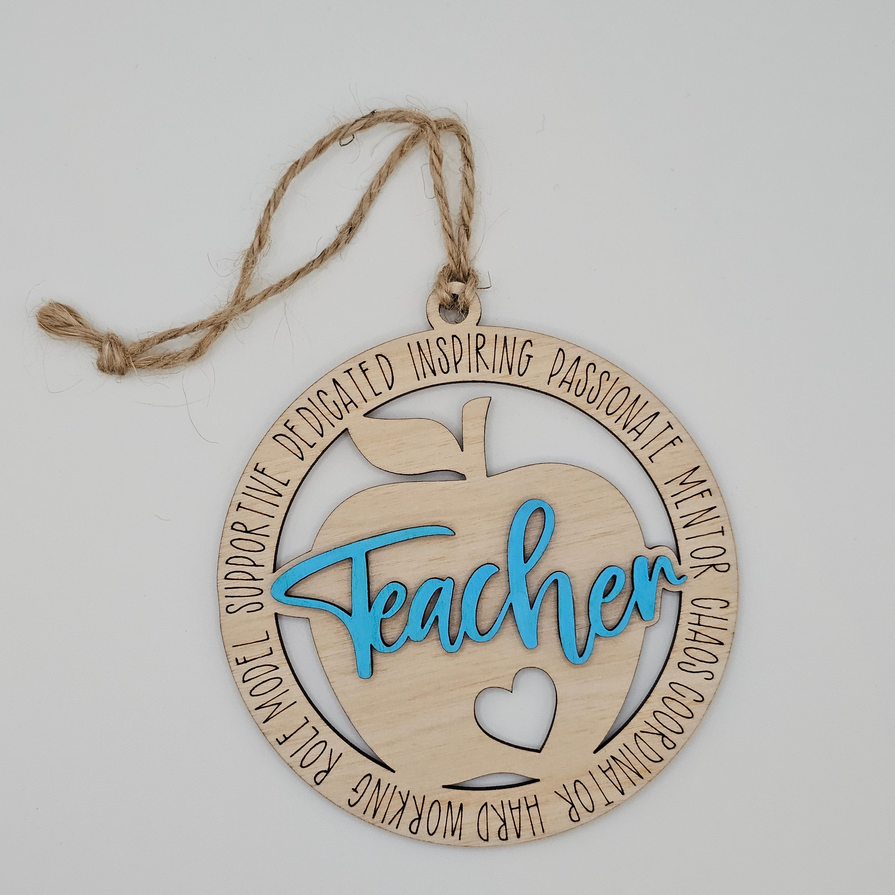 Layered laser cut Teacher apple wood ornament with turquoise painted script overlay, circular frame engraved with teacher descriptors, heart cutout at center, hung with natural jute cord