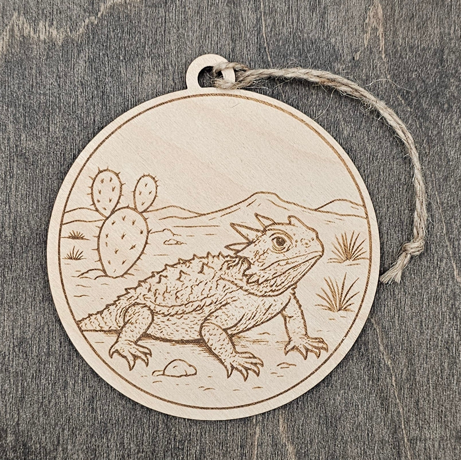 Laser engraved Texas Horned Lizard wood ornament, featuring a detailed illustration of this beloved Texas icon against a West Texas landscape with prickly pear cactus and mountains, hung with natural jute cord