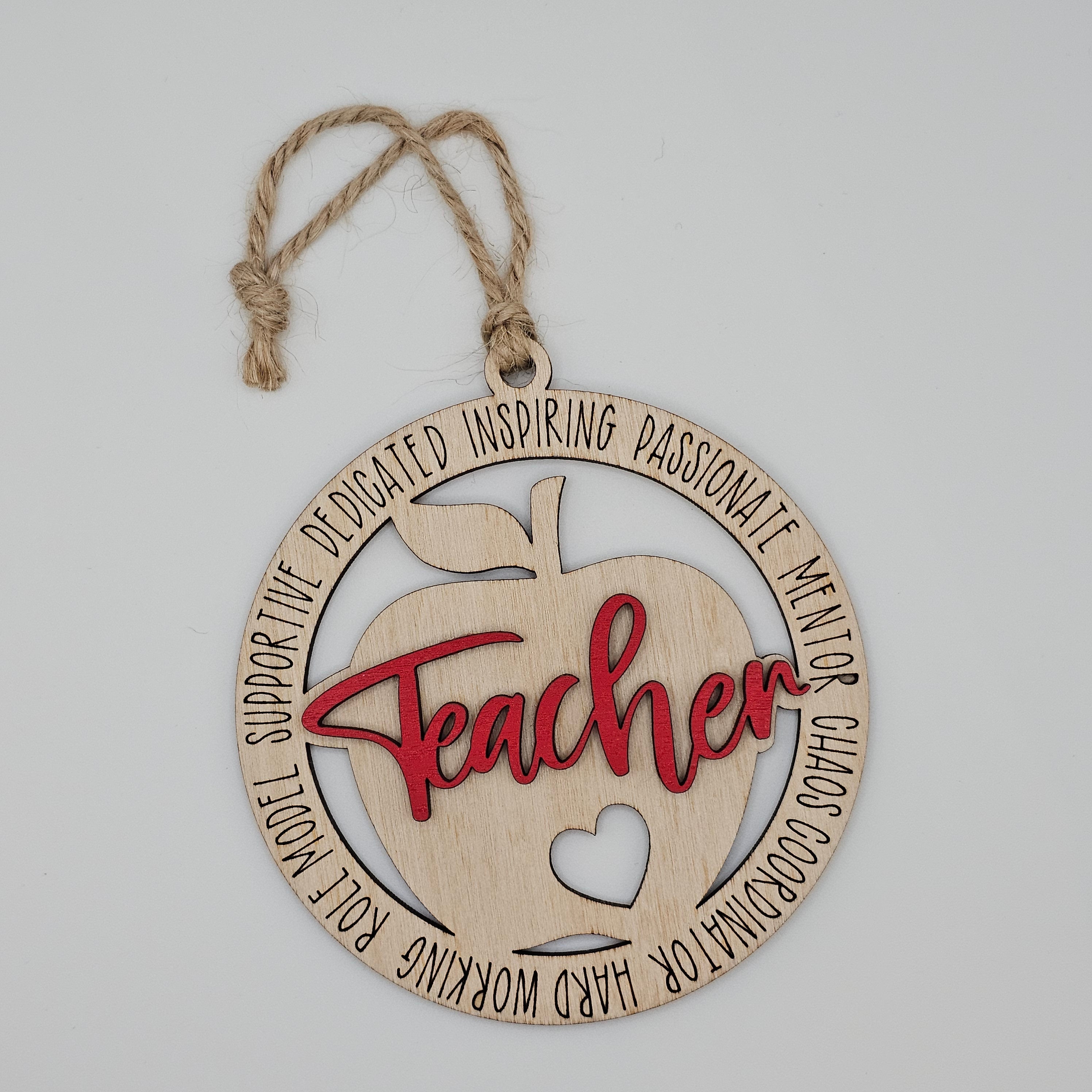 Layered laser cut Teacher apple wood ornament with red painted script overlay, circular frame engraved with teacher descriptors, heart cutout at center, hung with natural jute cord
