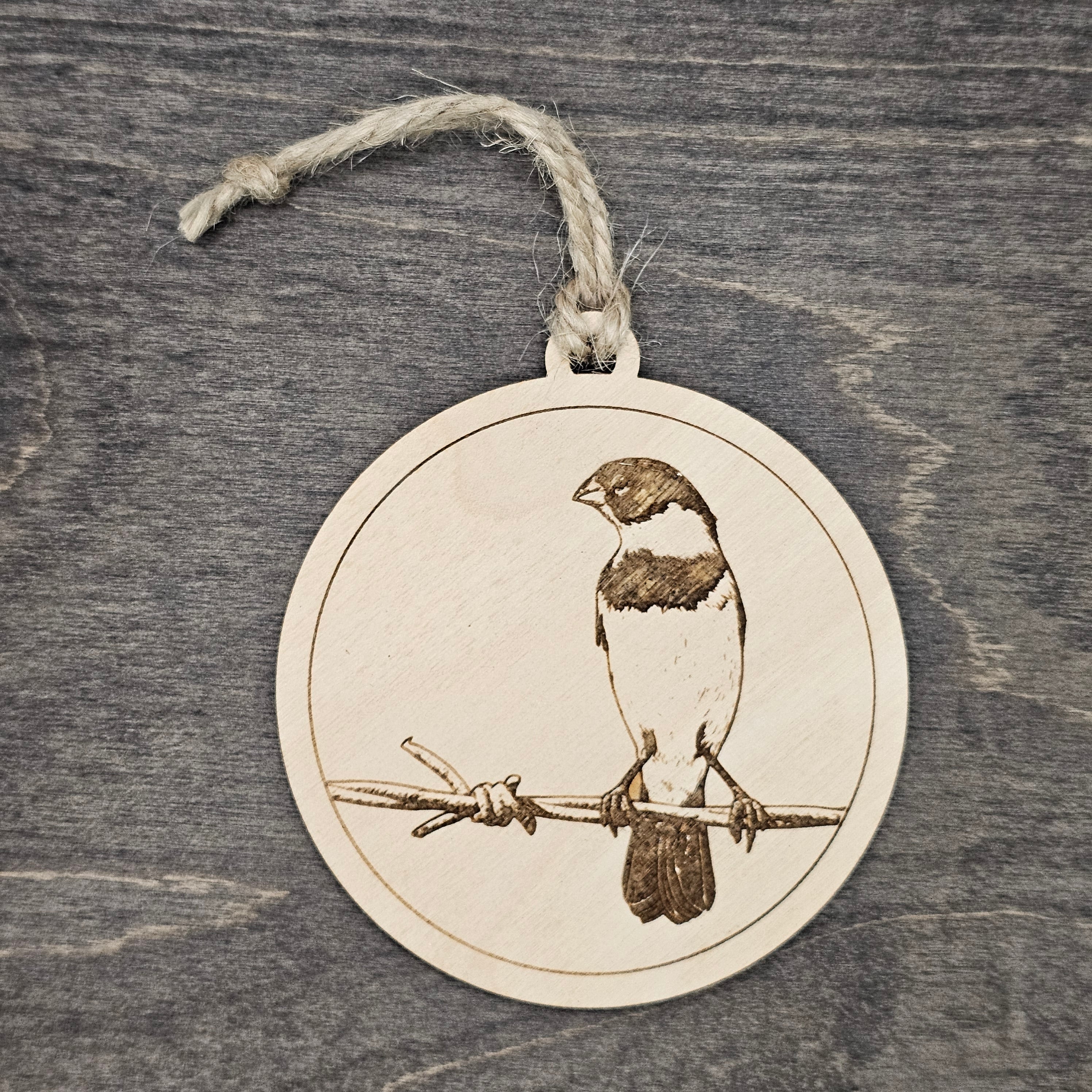 Laser engraved Morelet's Seedeater wood ornament, featuring a detailed bird illustration perched on a barbed wire fence, hung with natural jute cord